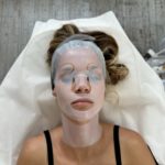 A relaxed woman with a bio-cellulose facial mask, lying down in a spa setting, showcasing a beauty treatment for skin rejuvenation.