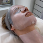 Woman receiving a professional skincare treatment with a bio-cellulose mask on her face and neck, lying in a clinic with a serene expression.