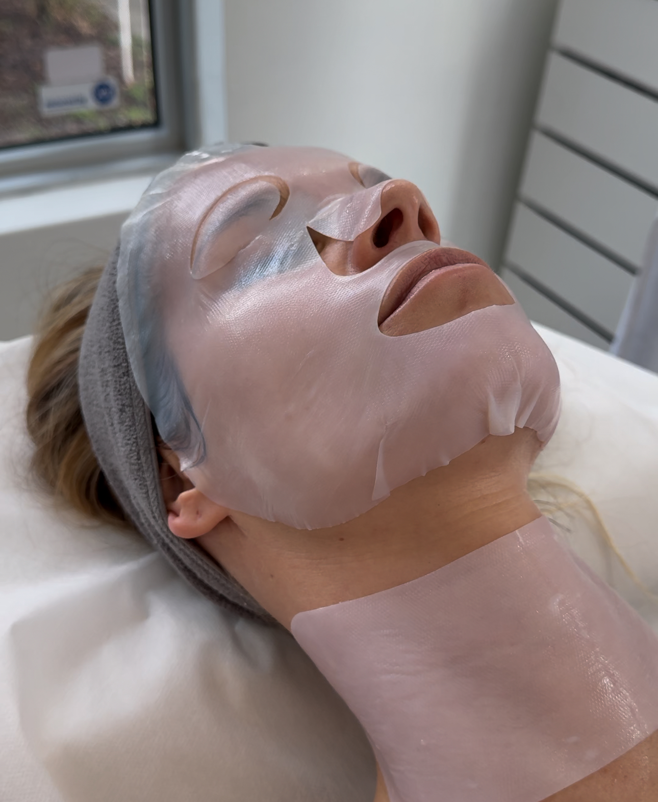 Woman receiving a professional skincare treatment with a bio-cellulose mask on her face and neck, lying in a clinic with a serene expression.