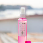 Bottle of Dermotechnology Bio-Lift Mist with a vibrant pink hue and a fresh flower on a stone ledge against a soft-focus waterscape