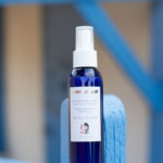 Bottle of Dermotechnology Bio-Immortelle Mist on a blue wooden bench with architectural elements in the background
