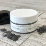 Closed jar of Dermotechnology Charcoal Clay Mask with raw charcoal pieces on a rustic wooden surface