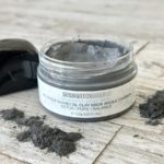 Open jar of Dermotechnology Charcoal Clay Mask with scattered charcoal pieces on a wooden background