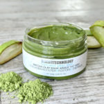Open jar of Dermotechnology Matcha Clay Mask with matcha powder and aloe slices on a wooden background