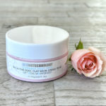 Closed jar of Dermotechnology Bio Active Rose Clay Mask next to a blooming pink rose on a wooden texture