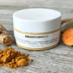 Closed jar of Dermotechnology Turmeric Clay Mask with turmeric powder and root on a textured wooden background