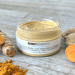 Open jar of Dermotechnology Turmeric Clay Mask with turmeric powder and root on a wooden surface
